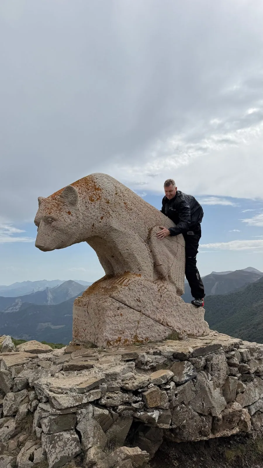 Rider climbing on a large stone bear sculpture on a mountain summit with peaks behind