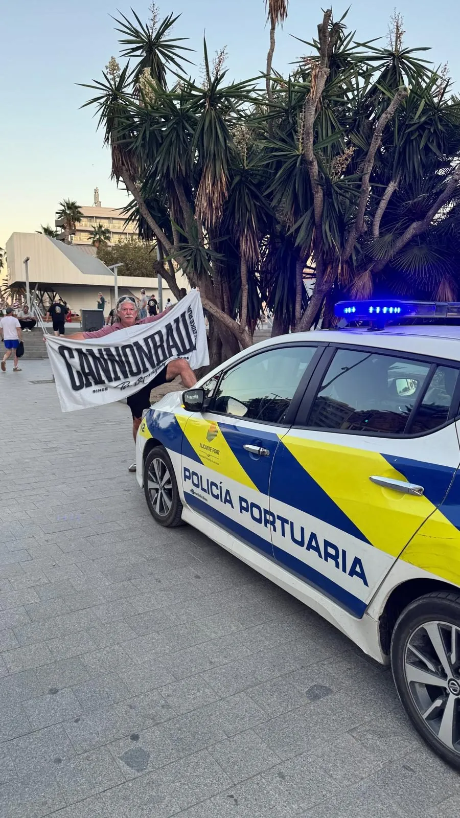 Rider holding a Cannonball banner next to a port police car on the Alicante promenade