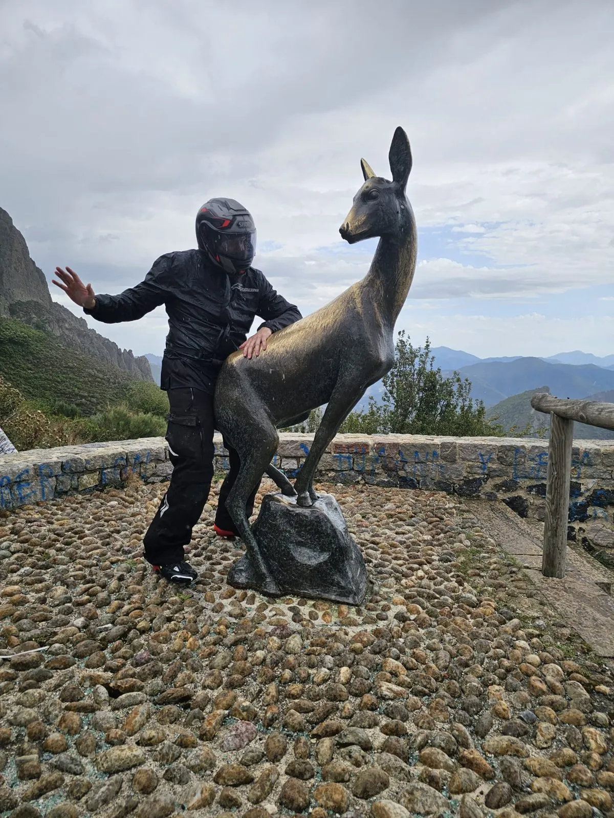 Rider in wet waterproofs posing beside a bronze deer statue at a misty mountain viewpoint