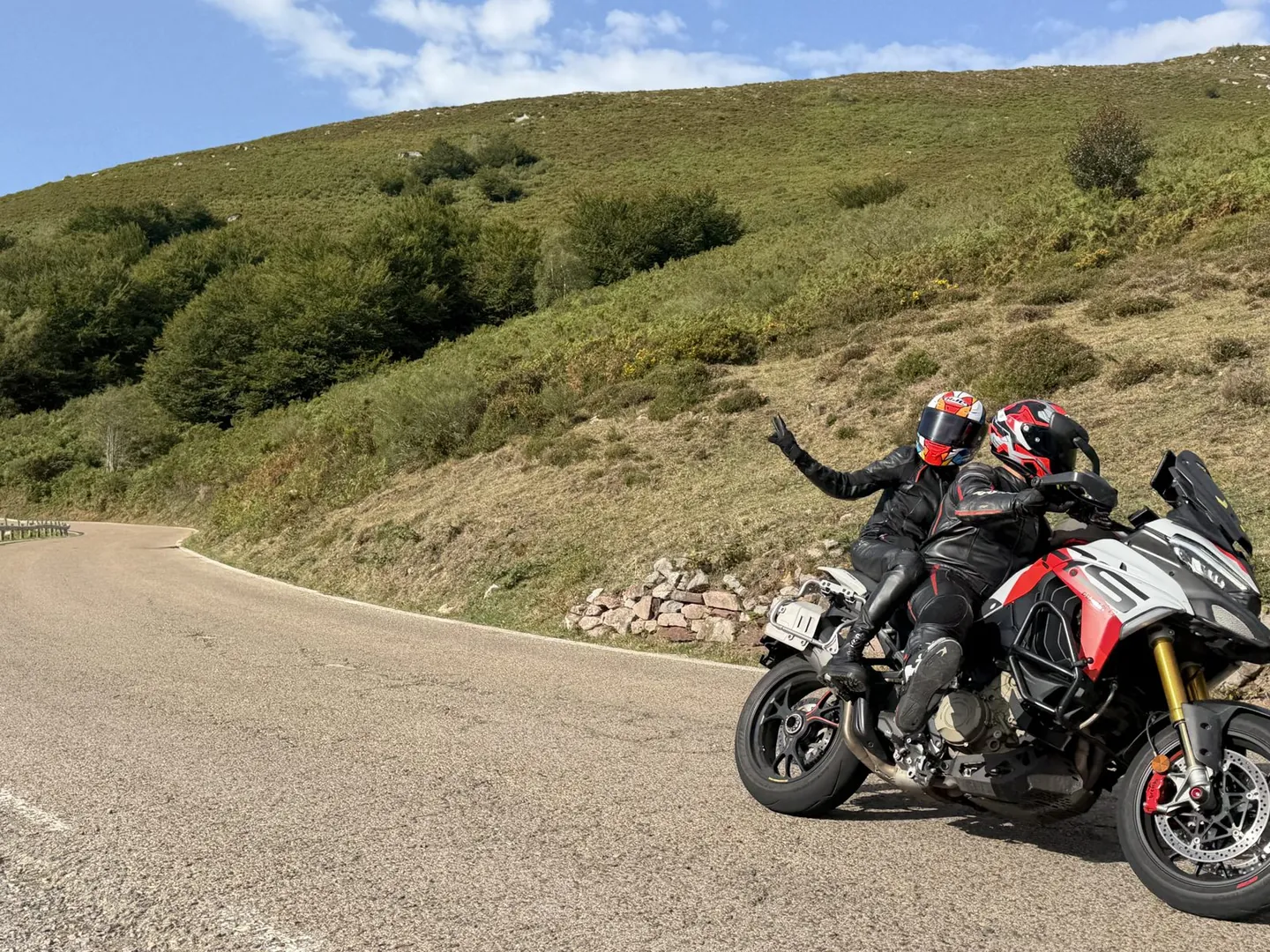 Two riders on a Ducati Multistrada waving on an empty mountain road in bright sunshine