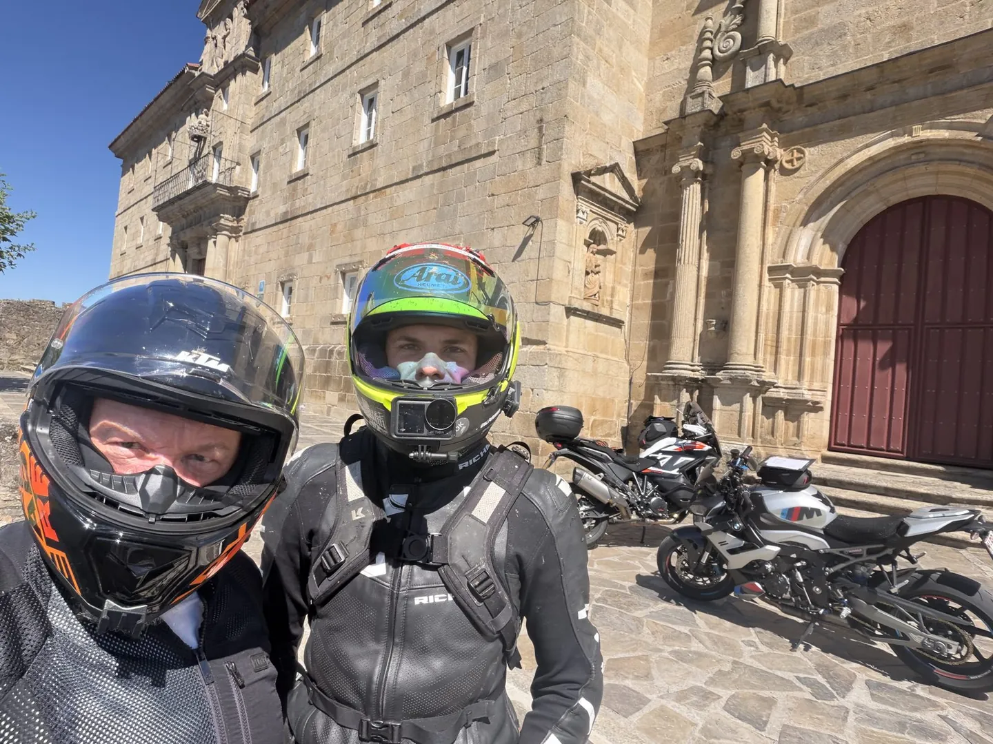 Two riders in helmets outside a large stone monastery with motorcycles parked by the entrance
