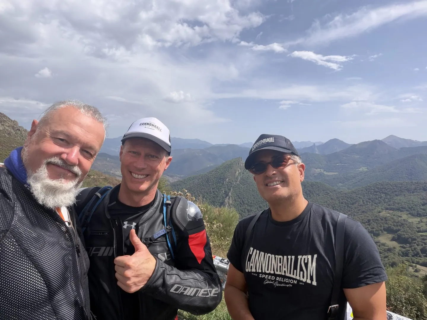 Three smiling riders in Cannonball caps and shirts at a mountain viewpoint