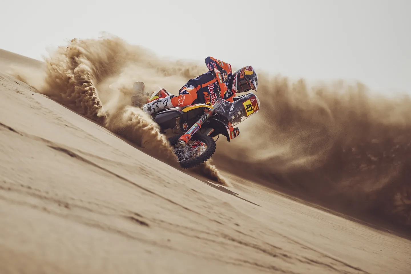 Red Bull KTM Factory Racing rider number 47 carving across a sand dune with a huge spray of sand