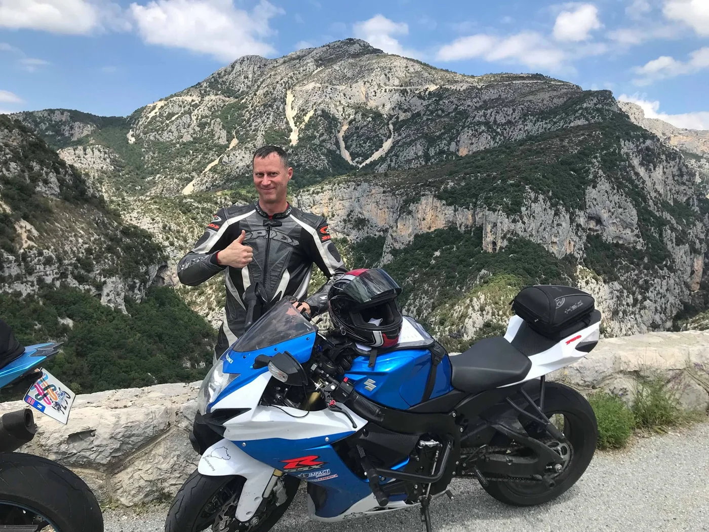 Jeremy Simmons giving a thumbs up beside his blue and white Suzuki GSX-R750 on a mountain road