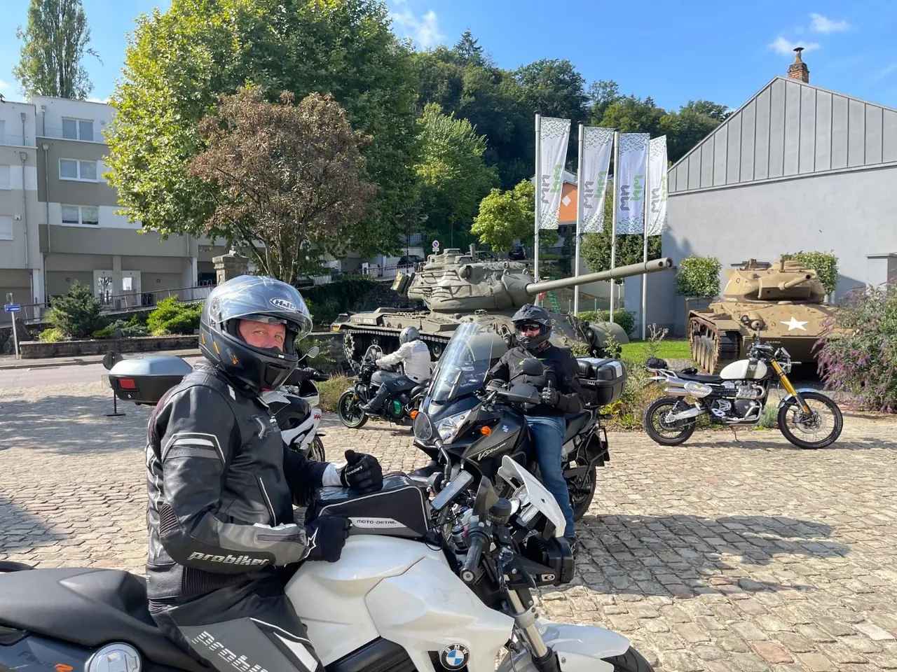 Riders regrouping on cobbles beside two preserved tanks on the Ardennes Tank Tour