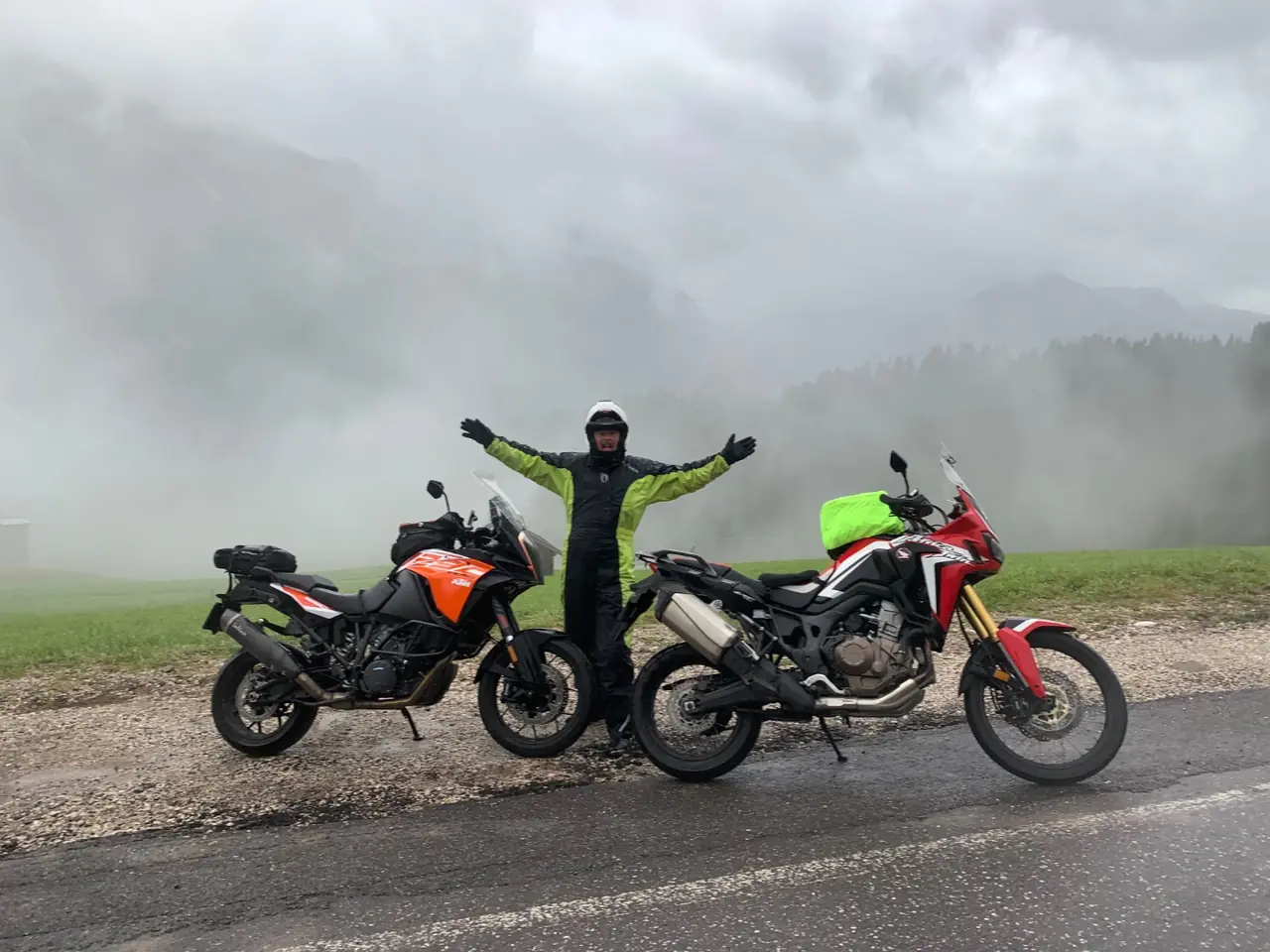 A rider in a hi-vis rain suit standing between two adventure motorcycles on a wet pass road, thick cloud filling the valley behind