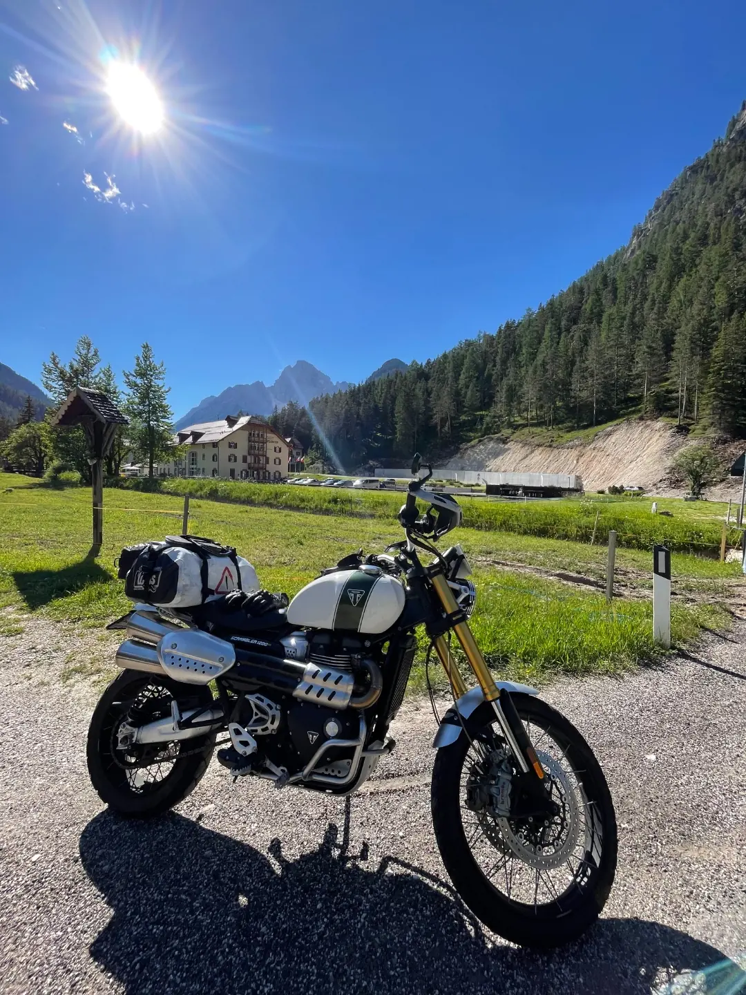 A Triumph Scrambler 1200 with soft luggage parked on gravel beside an alpine meadow, a village and jagged Dolomite peaks behind