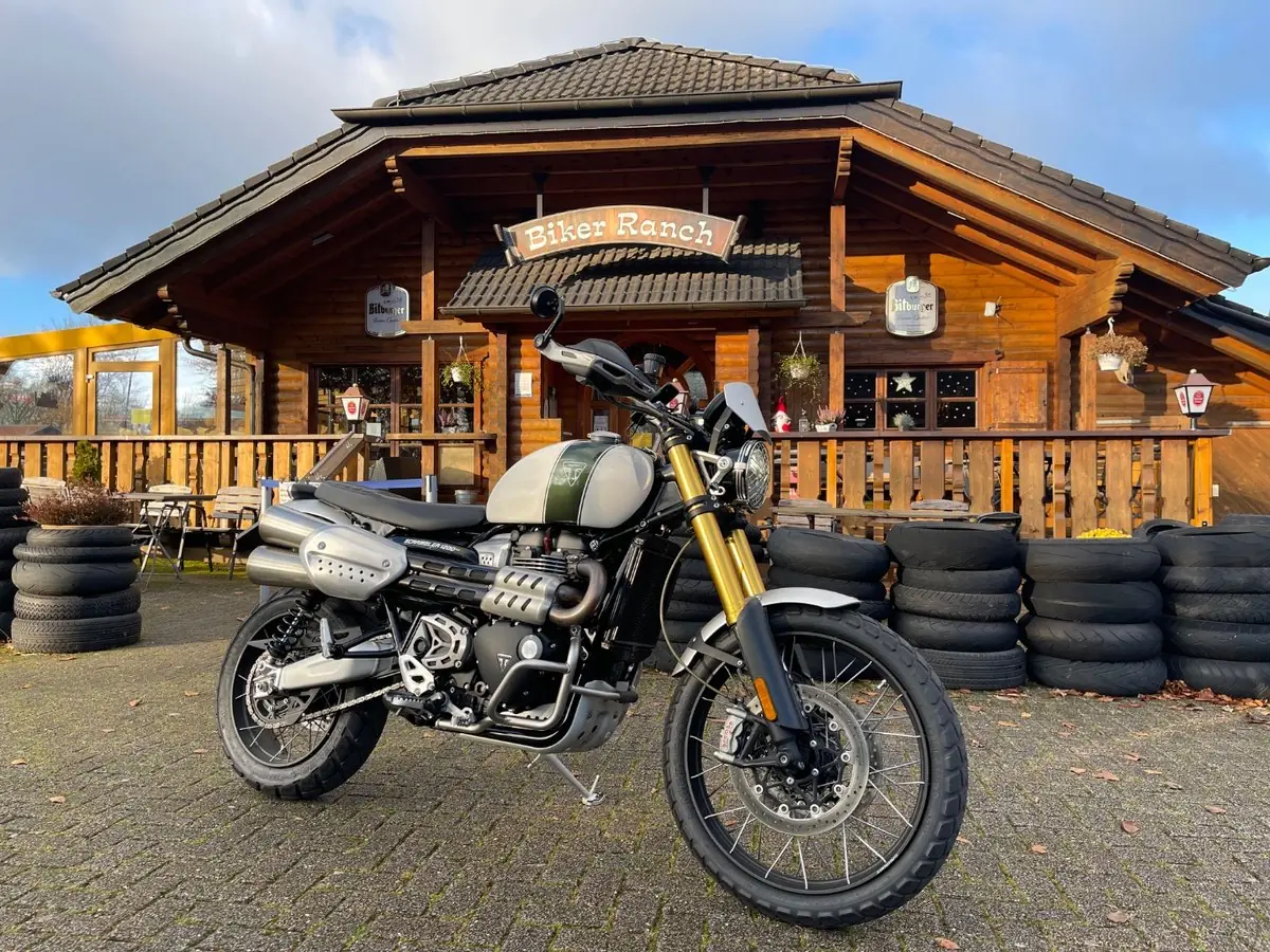 A Triumph Scrambler 1200 parked in front of the log cabin frontage of the Biker Ranch in Simmerath-Strauch, with stacks of tyres along the terrace