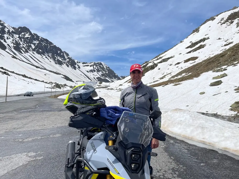 A rider standing beside a loaded Suzuki V-Strom adventure motorcycle at an Alpine pass summit with snowfields and a bare rock ridge behind
