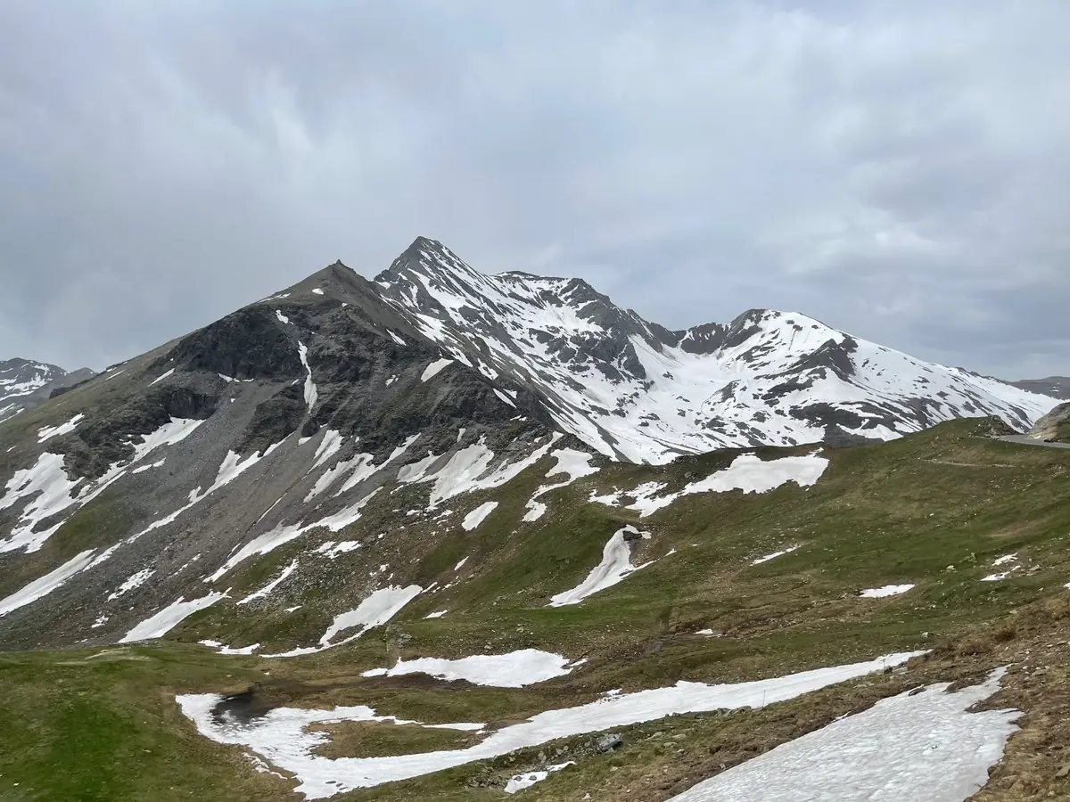 Grey rock peaks streaked with old snow above green alpine pasture on the Grossglockner High Alpine Road, with the road just visible traversing the slope on the right under a heavy overcast sky