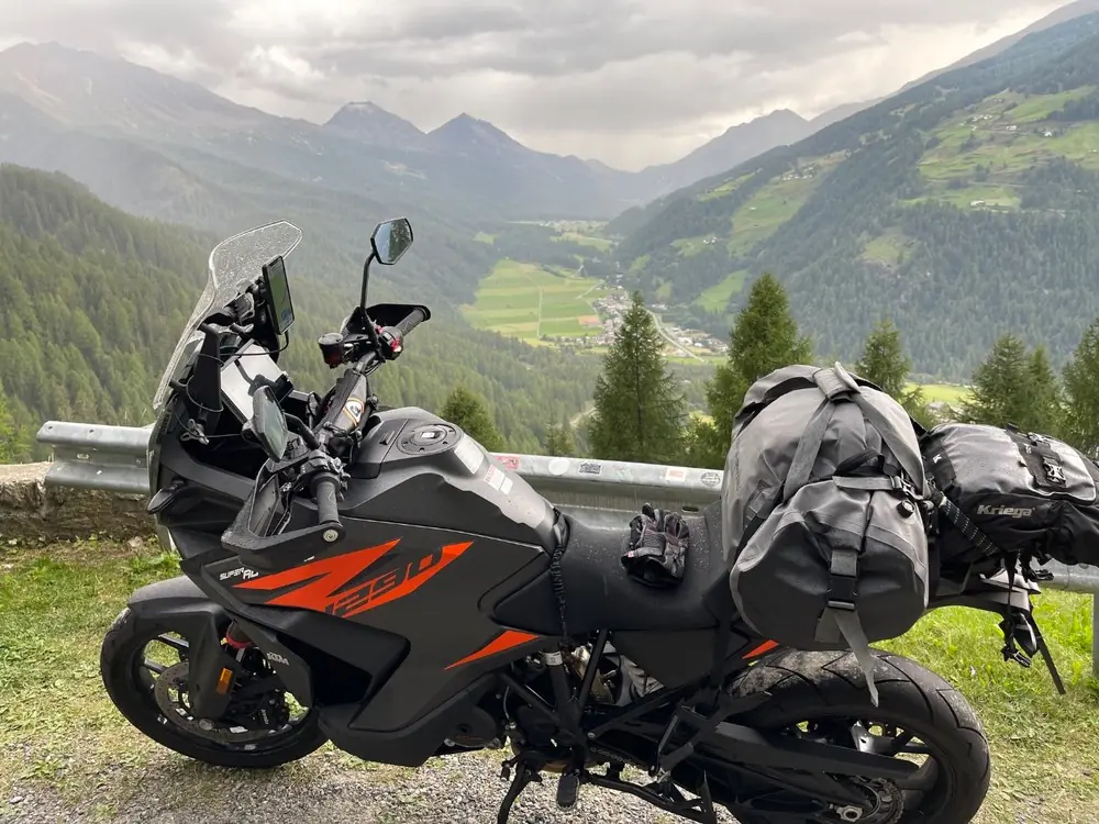A KTM 1290 Super Adventure loaded with soft luggage parked at a guardrail viewpoint above a broad green Alpine valley with a village and scattered farms below