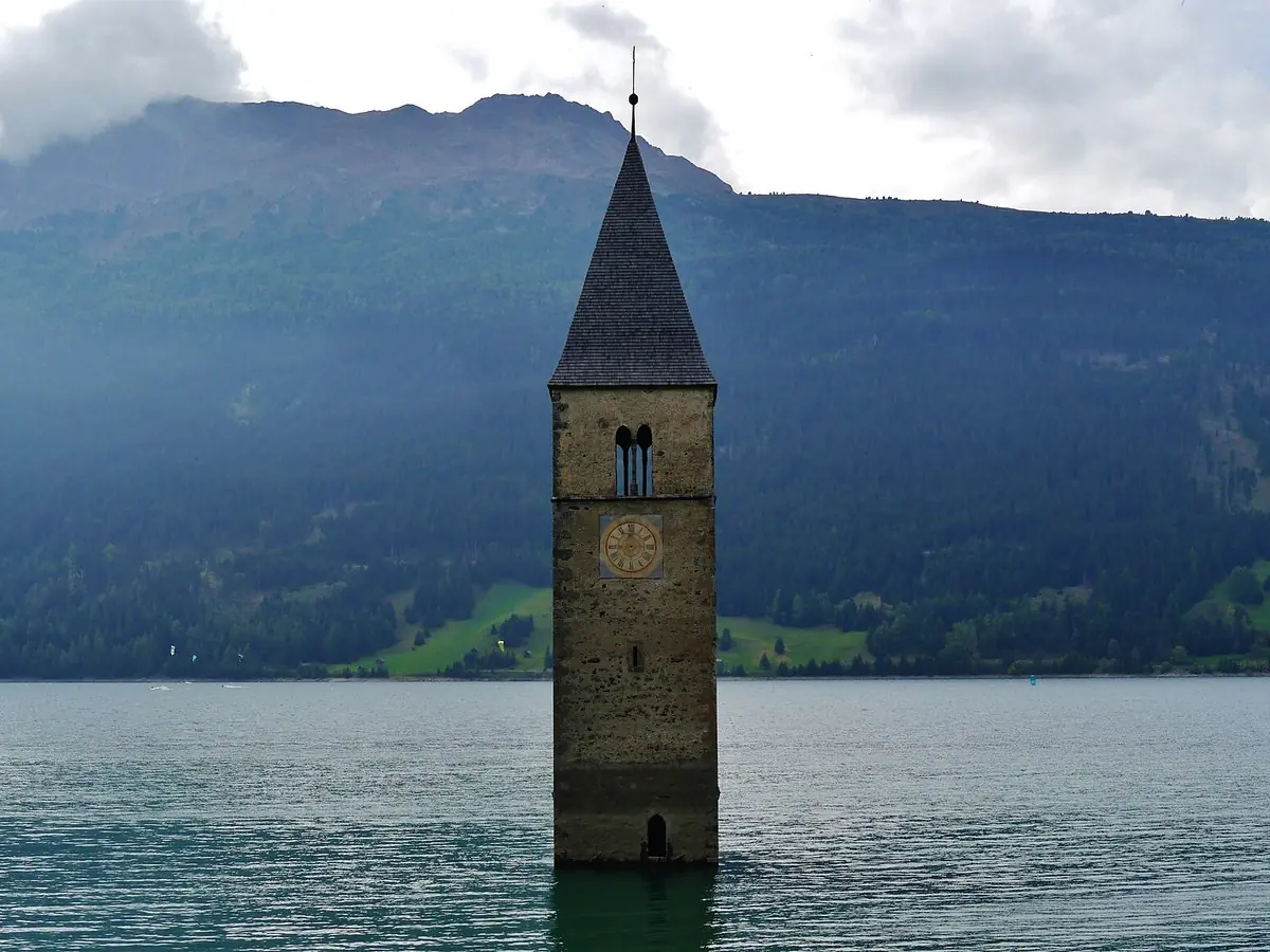 The stone bell tower of the drowned village of Graun standing alone in the turquoise water of the Reschensee reservoir, with forested slopes and cloud on the mountains behind