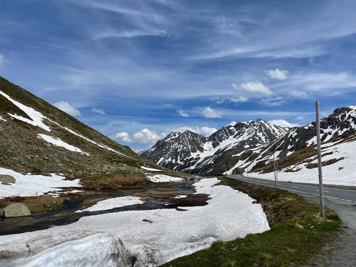 An open Alpine pass road running between deep banks of old snow and a meltwater stream, with snow-streaked peaks ahead under a blue sky