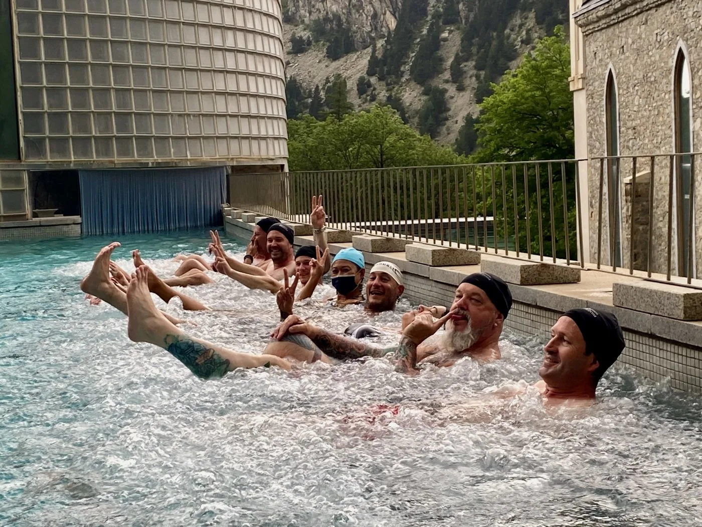 A group of riders in swim caps relaxing in a mountain spa pool, legs raised in the bubbles and making peace signs
