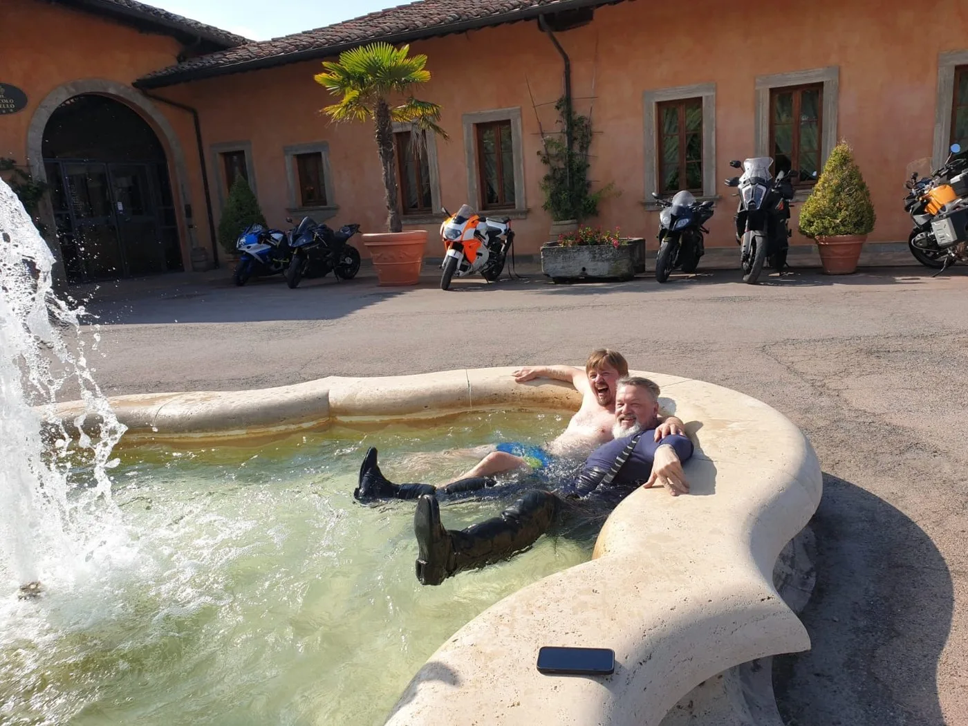 Alan, still in full riding gear and boots, and Petri in swim shorts, laughing as they sit in a hotel courtyard fountain with motorcycles parked behind them