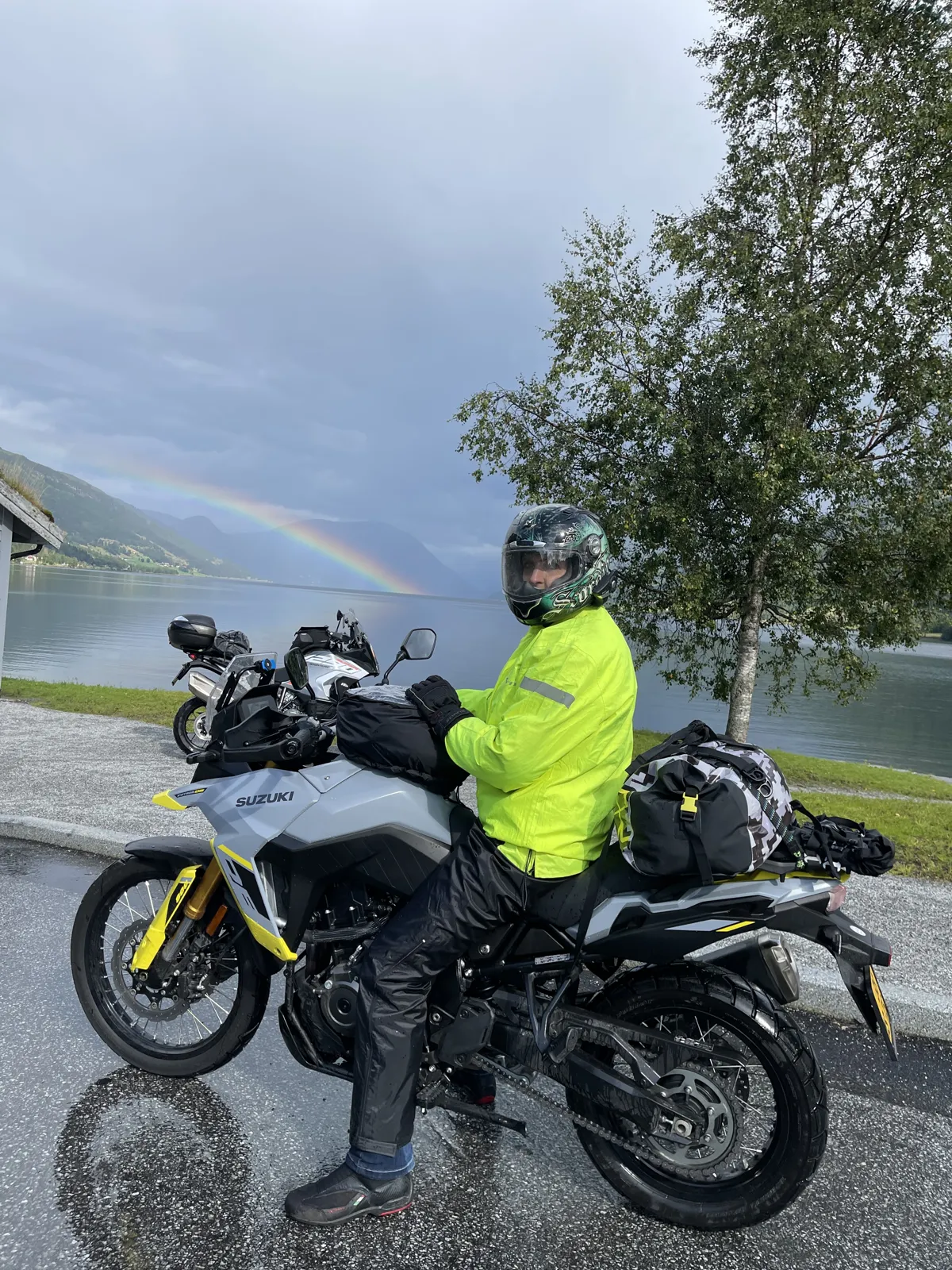Belinda in a fluorescent rain jacket on her grey Suzuki beside Jolstravatnet lake with a rainbow behind her