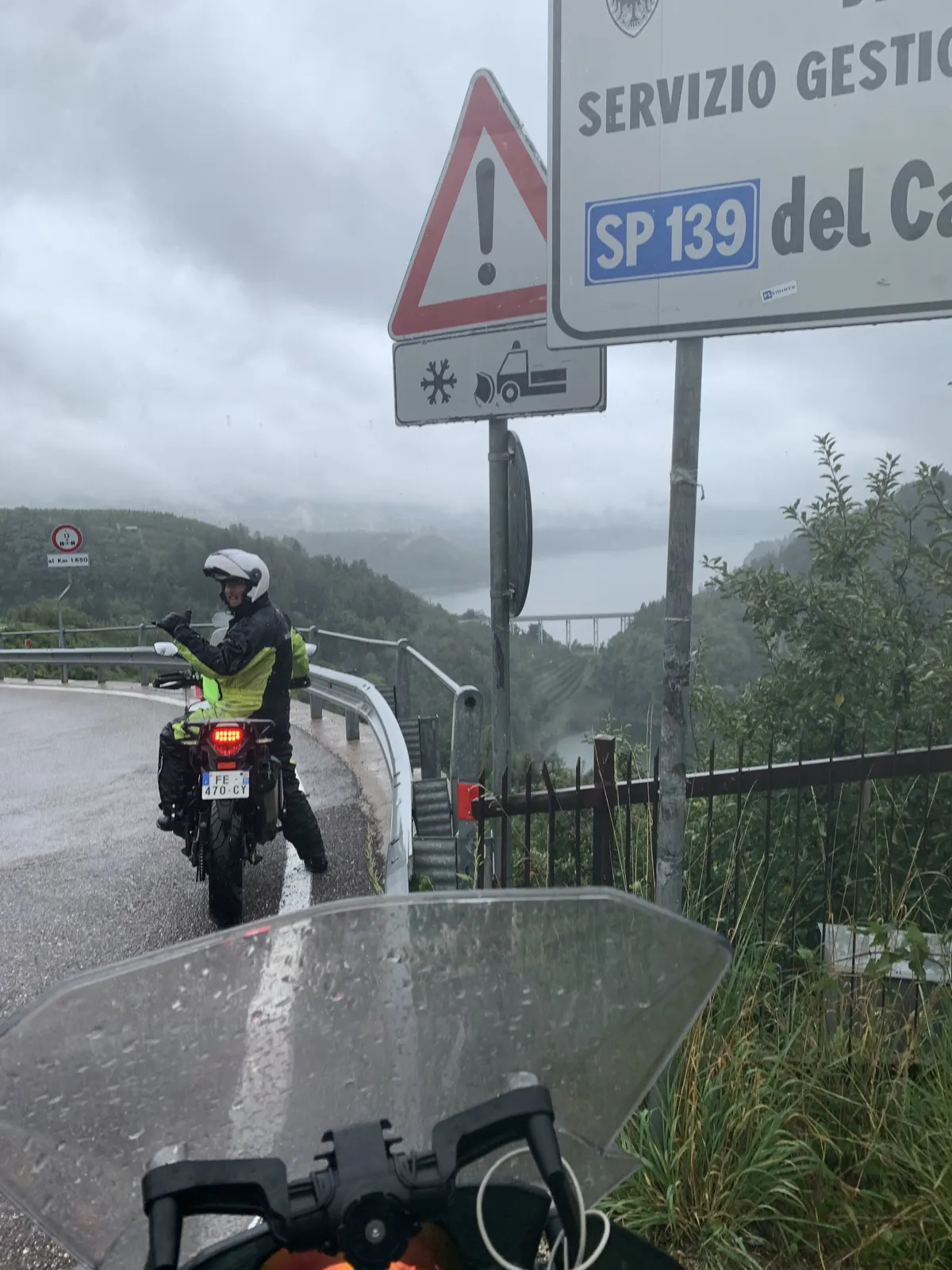 Rich in waterproofs beside his motorcycle next to an Italian road warning sign in rain and low cloud